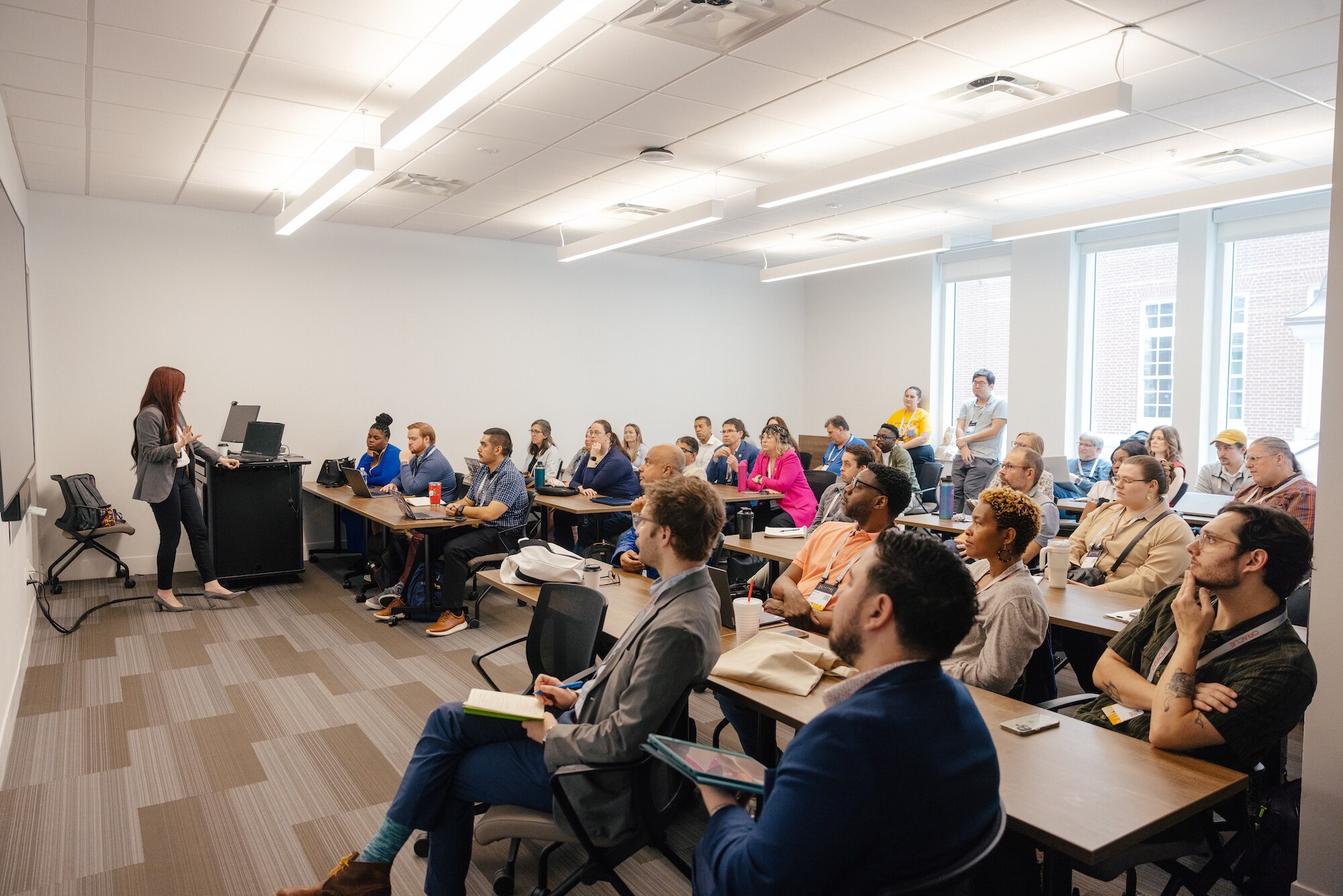 Cassandra Cardases presenting in 2026 stands at the front addressing a packed classroom-style room of attendees seated at desks with laptops, with a few people standing near the windows at the back.