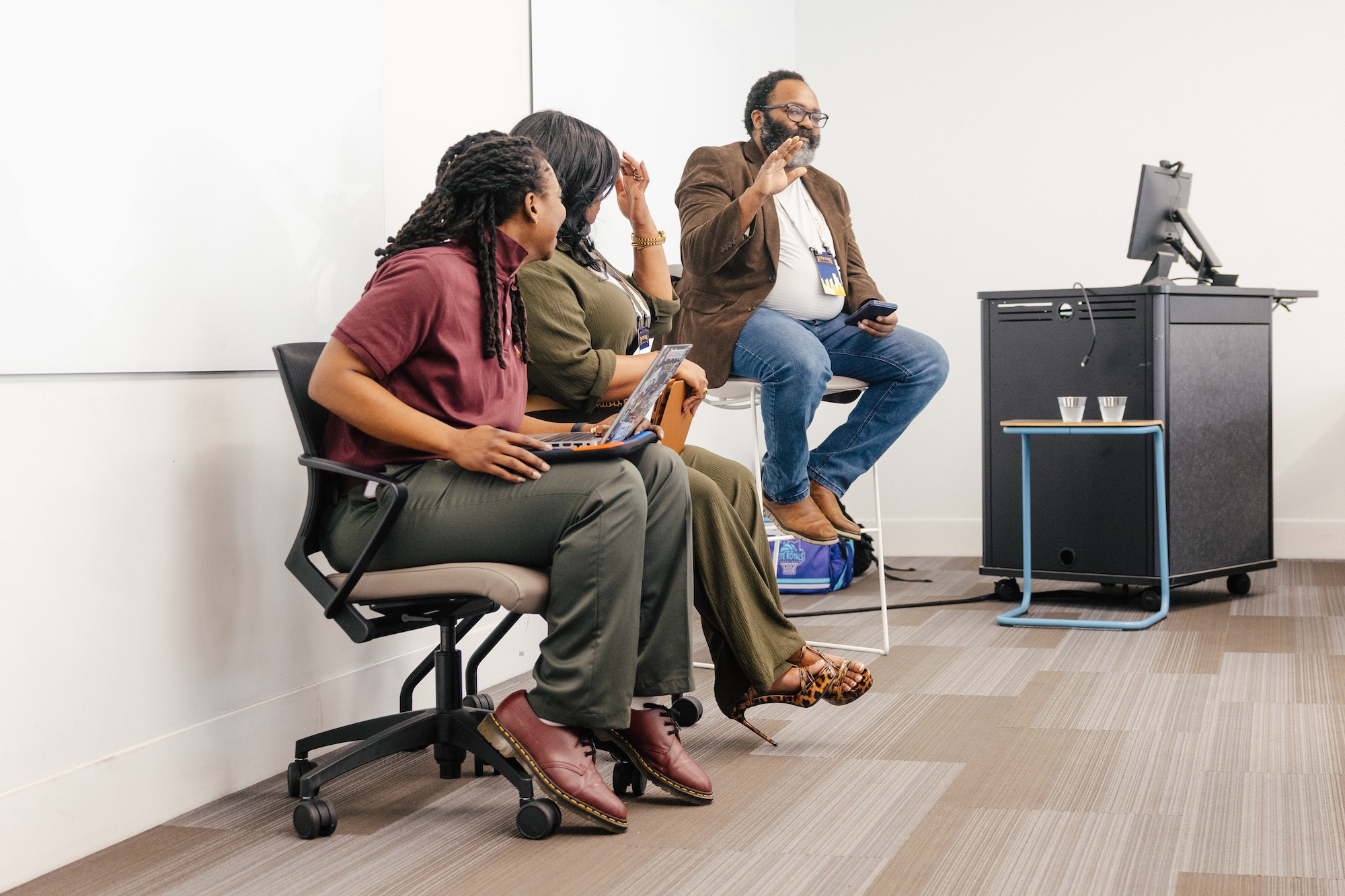 Jurnell Cockhren, Valencia Gooch, and Lena Winfree in a relaxed panel discussion, two seated with a laptop and one perched on a stool gesturing while speaking, with water glasses on a side table.